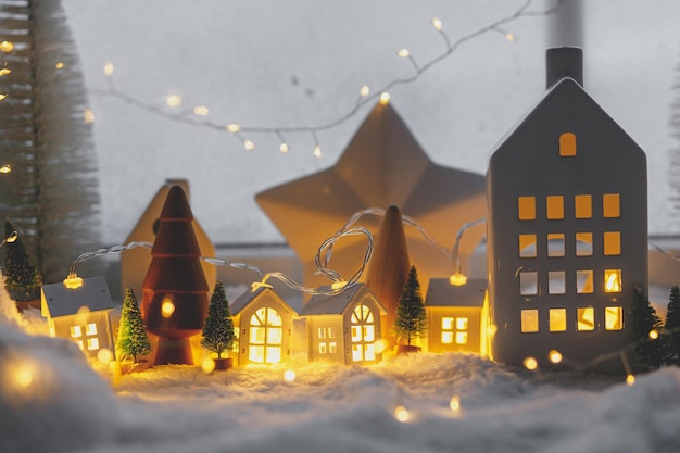 Cozy christmas miniature village Stylish little ceramic houses and wooden trees on soft snow blanket with glowing lights in evening Atmospheric winter village still life Merry Christmas