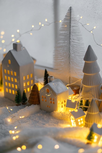 Cozy christmas miniature village Stylish little ceramic houses and wooden trees on soft snow blanket with glowing lights in evening Atmospheric winter village still life Merry Christmas