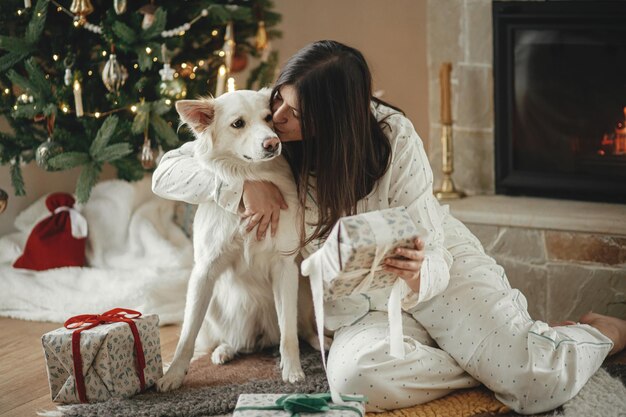 Photo cozy christmas morning beautiful woman in pajamas holding stylish christmas gift and hugging cute white dog at fireplace and decorated tree in festive living room merry christmas