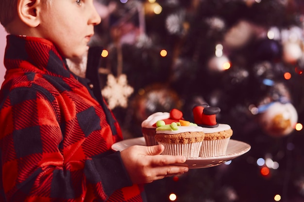 Cozy Christmas time Boy holds decorative festive cupcakes in front of the bokeh of Christmas tree Good holidays mood Selective focus