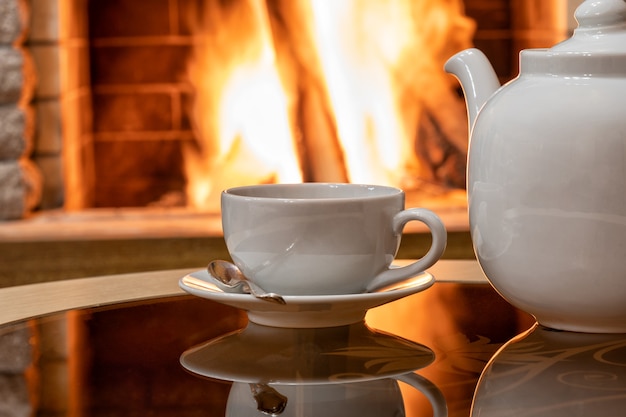 Cozy Fireplace scene and white cup of tea and teapot on a table, reflection from a fire on a glass table.