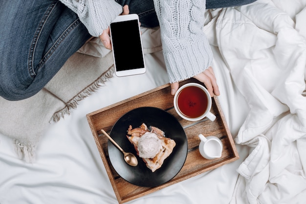 Accogliente flatlay del letto con vassoio in legno con torta di mele vegana, gelato e tè nero e donna in jeans e maglione grigio che tiene smartphone con copyspace nero su lenzuola bianche e coperte