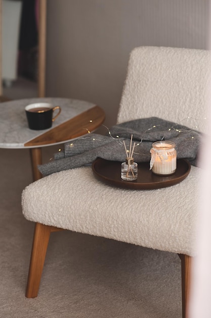 Cozy home atmosphere with liquid fragrance in glass bottle and\
burning scented candle with knit cloth textile on wooden tray on\
fluffy accent chair in bedroom