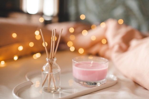 Photo cozy home atmosphere with pink scented candle on white ceramic tray and liquid diffuser in bed over glow christmas lights closeup winter holiday season