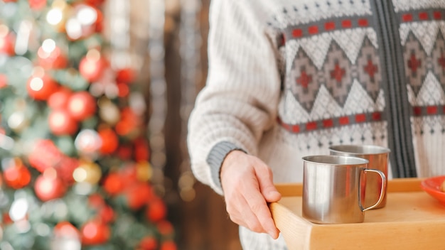 居心地の良いホームクリスマスパーティー。木製のトレイを使用して鋼のマグカップで温かい飲み物を提供する男のクロップドショット