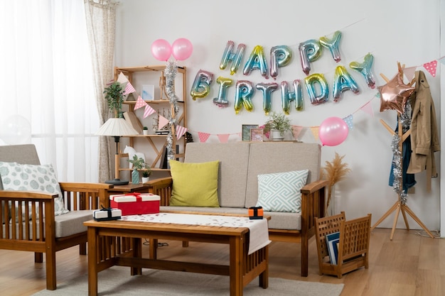 Photo cozy home interior with birthday party decorations: pink balloons, birthday garland on white wall and gifts on living room table.