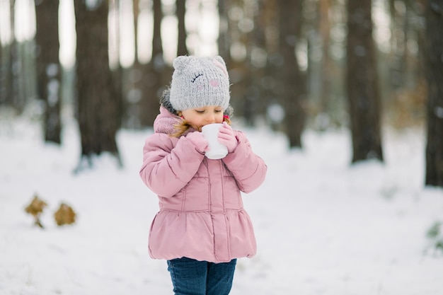 Idee accoglienti per picnic invernali all'aperto. la bambina si gode la bevanda calda invernale, in piedi tra gli alberi innevati invernali nella foresta. bevanda calda durante una passeggiata. copia spazio
