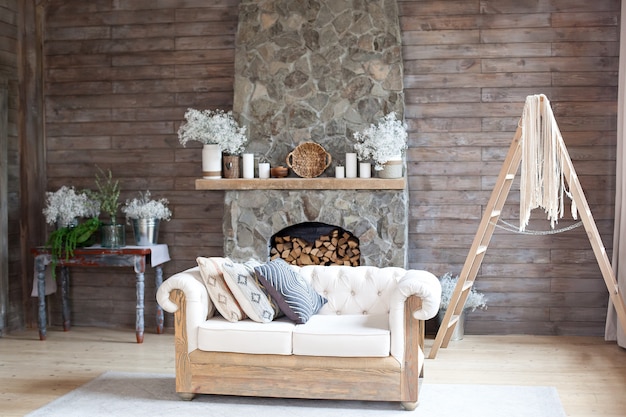 Cozy Interior living room white Sofa and Fireplace. Rustic Home Design for Warm Indoor Space Alpine Vacation. Modern Cottage Living Room Decor with Wood Wall and Furniture. Scandinavian style. Boho