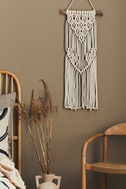 Cozy interior of stylish bedroom with design decoration, neutral macrame, dried flowers in vase , chair, beautiful bed sheets, blanket ,pillows and personal accessories