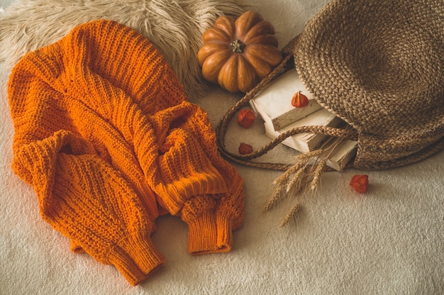 Cozy knitted warm orange sweater with old books and vintage straw bag on white warm plaid with pumpkin, physalis, Autumn books and reading. autumn mood. Autumn Winter time.