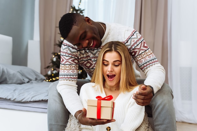 Cozy moments in winter holidays at decorated tree. Seasonal greetings concept. A young woman is surprised by a gift that a boyfriend gives her.