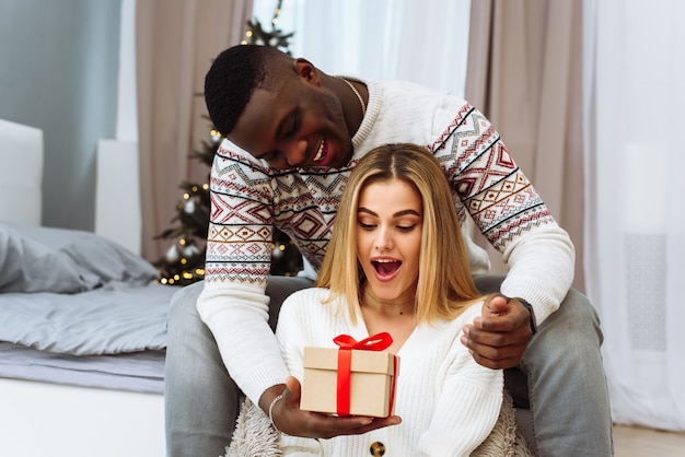 Momenti accoglienti durante le vacanze invernali all'albero decorato. concetto di saluti stagionali. una giovane donna è sorpresa da un regalo che le fa un fidanzato.