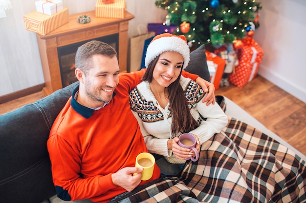 Cozy picture of happy young man and woman sitting on sofa and hold cups with hot drink. 