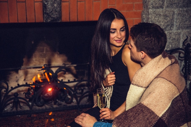 Cozy picture of young man and woman sit together and look at each othr. She smiles. Guy wear blanket on shoulders. They sit together at fireplace