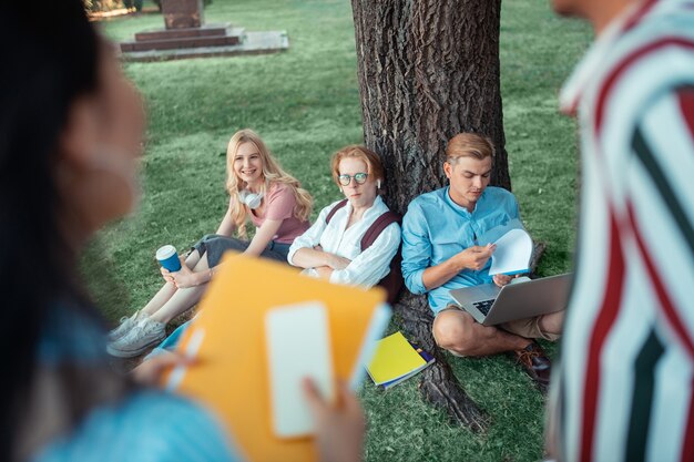 Foto posto accogliente. tre studenti concentrati seduti sotto il grande albero davanti ai loro compagni di gruppo e facendo i compiti.