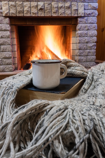 Фото Уютная сцена перед камином с белой эмалированной кружкой с чаем, книгой и шерстяным шарфом.