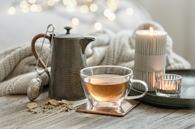 Photo cozy still life with a glass cup of tea, a teapot and candles on a blurred background with bokeh.