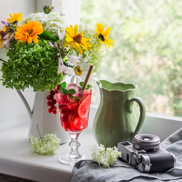 Cozy summer still life bouquet of flowers glass of lemonade jug\
vintage camera on the windowsill in a room