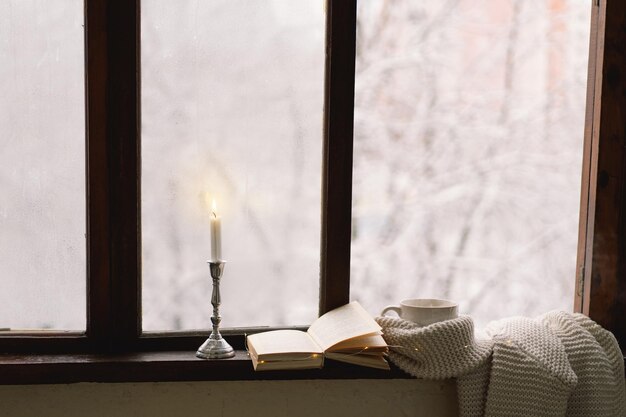 Cozy winter still life Cup of hot tea and an open book with a warm sweater on a vintage wooden windowsill Cozy home concept Sweet home