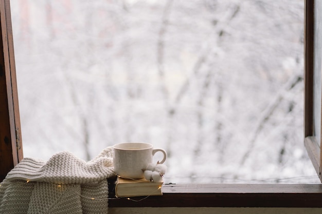 Cozy winter still life Cup of hot tea and an open book with a warm sweater on a vintage wooden windowsill Cozy home concept Sweet home