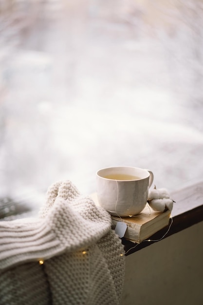 Photo cozy winter still life cup of hot tea and an open book with a warm sweater on a vintage wooden windowsill cozy home concept sweet home