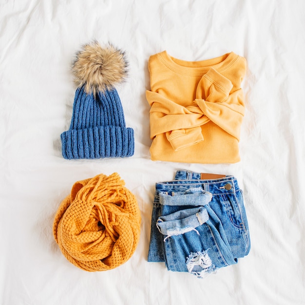 아늑한 노란색 스웨터, 청바지, 스카프, 모자는 흰색 시트에 침대에 있습니다. 여성의 세련된 가을 또는 겨울 복장. 트렌디한 옷 콜라주. 평평한 평지, 평면도.
