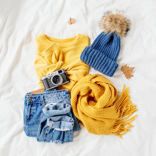 Cozy yellow sweater, blue jeans, scarf and hat on bed on white sheet. Women's stylish autumn or winter outfit. Trendy  clothes collage. Flat lay, top view.