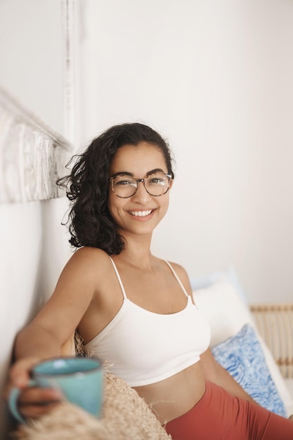 Cozyiness wellbeing and people concept Charming young eastern hipster woman in glasses white bra and leggings drink fresh coffee from mug smiling camera happily having rest after work