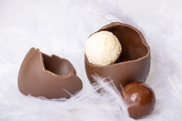 Cracked chocolate easter egg with chocolate small round candies and empty egg shell in nest of white feathers