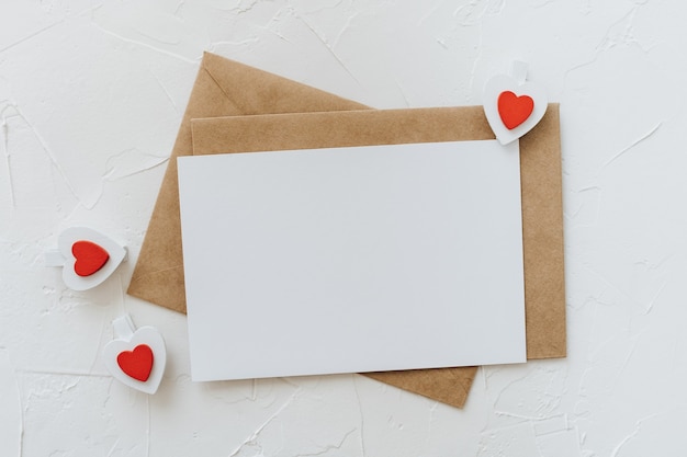 Craft envelopes, white blank sheet of paper and wooden clips hearts on a white background. Valentine's day concept.