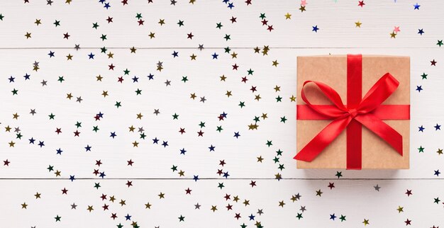 Ремесленная подарочная коробка с красной лентой на белом дереве с конфетти и копией пространства, вид сверху