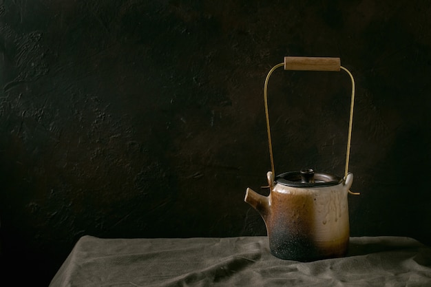 Craft handmade ceramic teapot kettle with brass handle for tea ceremony standing on linen tablecloth in dark room.