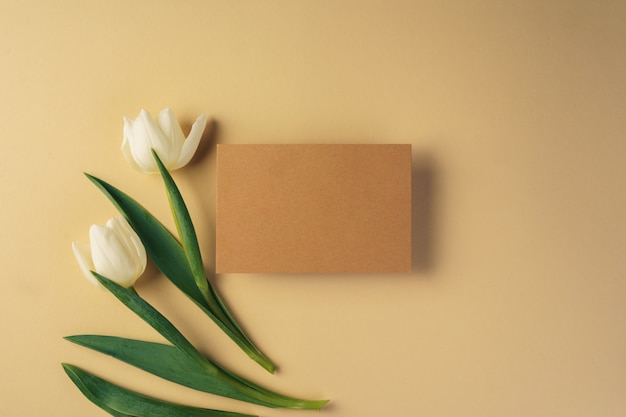 Craft paper card surrounded by fresh tulips on beige surface