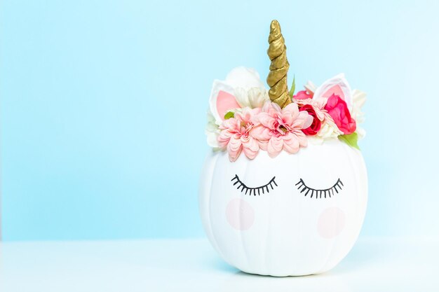 Craft pumpkin painted white and decorated with pink flowers as unicorn on blue background.