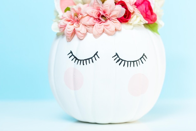 Craft pumpkin painted white and decorated with pink flowers as unicorn on blue background.