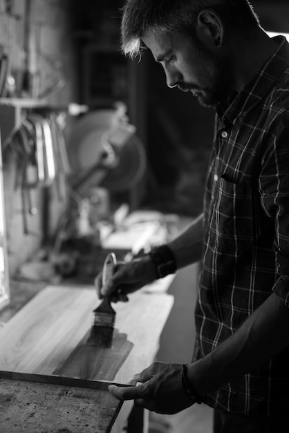 Photo craftsman applies varnish on wooden board by paintbrush in his carpentry workshop