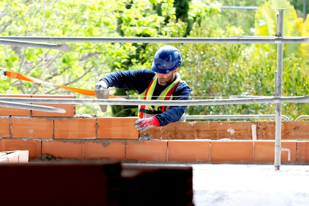 A craftsman in a helmet wearing a safety belt applies a cement mixture with a spatula to a brick