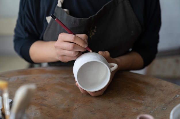 Craftswoman makes sketch of future pattern on white mug to decorate kitchen with clay dish