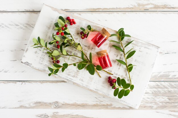 Cranberry essential oil in glass bottles with a cork lie on a white wooden stand with branches of natural cranberries. Selective focus.