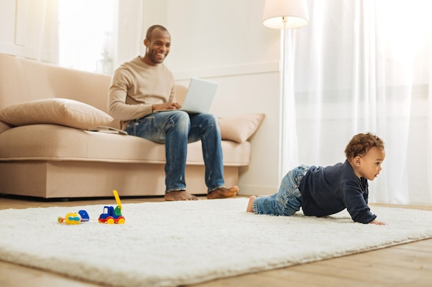 Strisciando. padre afroamericano sorridente e giovane premuroso che lavora al suo computer mentre era seduto sul divano e guardava il suo piccolo figlio che gattonava