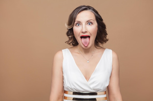 Crazy adult woman with big eyes tongue out . Emotional expressing woman in white dress, red lips and dark curly hairstyle. Studio shot, indoor, isolated on beige or light brown background