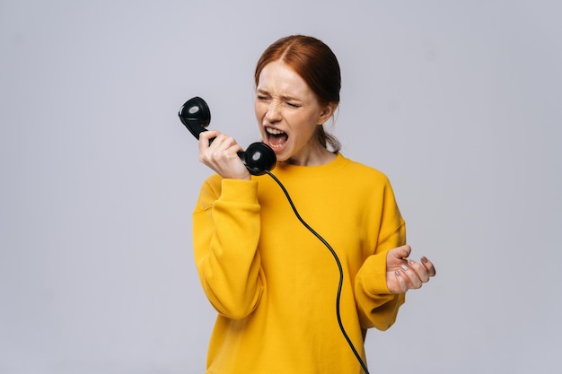 Crazy angry young woman in stylish yellow sweater talking on retro phone and screaming in handset