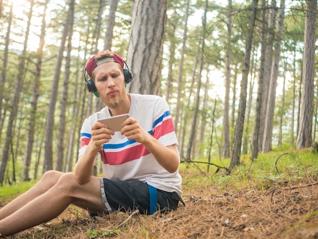 Crazy bearded hipster in casual and cap sit in the nature forest with phone and headphones playing gaming on smartphone