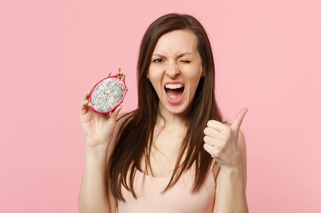 Crazy blinking young woman showing thumb up, holding half of fresh ripe pitahaya, dragon fruit isolated on pink pastel background. People vivid lifestyle relax vacation concept. Mock up copy space.