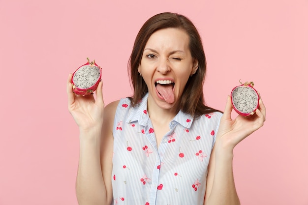 Crazy blinking young woman showing tongue holding halfs of fresh ripe pitahaya, dragon fruit isolated on pink pastel wall background. People vivid lifestyle relax vacation concept. Mock up copy space.