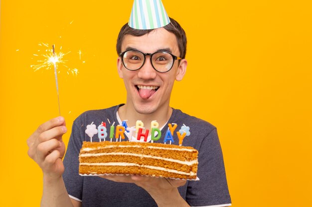 Crazy cheerful young guy with glasses holding a burning candle in his hands and a congratulatory cake on a yellow space. birthday and anniversary celebration concept. advertising space