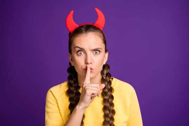 crazy childish lady holding finger on lips frightening evil facial expression wear halloween red horns casual yellow pullover isolated purple color wall