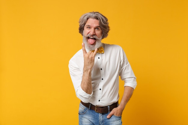 Crazy elderly gray-haired mustache bearded man in shirt and bow tie posing isolated on yellow background. People lifestyle concept. Mock up copy space. Showing tongue, depicting heavy metal rock sign.