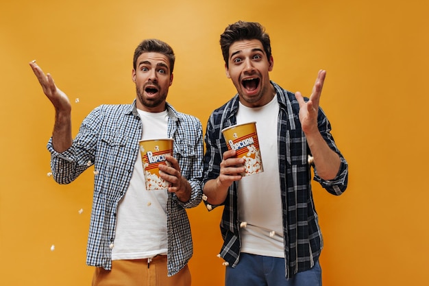Crazy emotional young brunet men in white tshirts and checkered shirts look surprised hold popcorn and pose on orange background