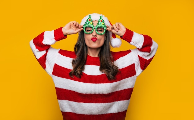 Crazy and excited beautiful modern woman in funny Christmas glasses and Santa hat is having fun while posing against a yellow wall